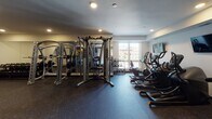 Located at 1732–1798 First Ave S in Minneapolis, MN, this fitness center is a well-appointed, single-floor facility designed to support a full range of workout routines. The space is organized into distinct functional zones, each outfitted with professional-grade equipment. The cardio area features treadmills and elliptical machines positioned alongside wall-mounted televisions for an engaging workout experience. Natural light filters in through a window, offering views of the surrounding Minneapolis streetscape. The strength training zone is equipped with a Hoist cable machine system, a squat/power rack, and a comprehensive dumbbell rack that lines a full-length wall mirror, allowing users to monitor their form throughout their session. The rubber-floored surface throughout provides both safety and durability. A dedicated functional training area rounds out the facility, stocked with stability balls, medicine balls, resistance bands, and yoga mats organized on a storage rack. This corner of the gym is distinguished by a bold, large-scale mural depicting a dynamic athletic figure set against a vivid geometric background, adding an energizing artistic dimension to the space. A water fountain and sanitation station are conveniently located nearby. Recessed lighting throughout keeps the environment bright and focused, contributing to an overall atmosphere that is both motivating and practical.