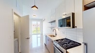 This single-floor unit showcases a clean, modern design characterized by polished concrete floors that run throughout the space, reflecting natural light and lending an industrial-chic aesthetic. The living area is a standout feature, anchored by expansive floor-to-ceiling corner windows that frame sweeping urban views of the surrounding cityscape, including neighboring buildings and tree-lined streets below. Multiple windows across the unit ensure abundant natural light in every room.

The kitchen is well-appointed with flat-front cabinetry in a warm wood tone complemented by crisp white upper cabinets and subway tile backsplash. Stainless steel appliances, including a gas range and built-in microwave, are paired with a matte black faucet and pendant lighting, creating a contemporary and functional cooking space. A tall window in the kitchen further enhances the bright, airy atmosphere.

The bedrooms are generously proportioned, each featuring large windows with urban views, polished concrete flooring, wall-mounted ductless HVAC units for individual climate control, and built-in closets with sliding or hinged doors. The bathroom is cleanly finished in white with a toilet and vanity. Throughout, the unit maintains a consistent, minimalist palette of white walls and neutral tones.