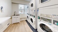 This 90 square foot laundry facility, located at 1200 Howie Ave in Coquitlam, BC, offers a compact yet efficient space for residents. The single-floor layout maximizes functionality within its limited area. Upon entering, you're greeted by a narrow hallway with light wood-look flooring and crisp white walls, creating a clean and bright atmosphere.

The main area houses multiple commercial-grade washers and dryers, arranged in two rows along the walls. These white appliances feature digital displays and coin-operated systems, catering to various laundry needs. A dedicated sink area with a modern faucet provides convenience for hand-washing delicates.

Natural light floods the space through a window, offering a view of the surrounding area and enhancing the room's openness. Practical features include informative posters on the walls, providing guidance on laundry best practices and facility rules. A wall-mounted dispenser and a trash receptacle add to the room's utility.

The space prioritizes function without sacrificing cleanliness and order. Its design reflects a thoughtful approach to communal laundry facilities, ensuring residents have access to essential amenities in a well-maintained environment.
