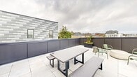 Located at 1406 Tacoma Ave S in Tacoma, Washington, this thoughtfully designed single-floor space spans a generous footprint across four distinct areas, offering a well-rounded mix of social, functional, and recreational environments.

The interior common areas are defined by clean white walls, light hardwood flooring, and striking black acoustic panel beams overhead that add both visual interest and sound management. Abundant natural light pours in through large windows, many of which frame elevated views of the surrounding Tacoma cityscape. Seating is arranged around natural wood communal tables paired with warm-toned bentwood chairs, creating an inviting atmosphere suited for collaboration or casual gathering.

A centrally positioned kitchen station features open shelving, a sink, and a preparation counter finished in calming blue-grey tones, serving the surrounding common areas efficiently.

A dedicated lounge and game room offers a more sophisticated ambiance, with rich wood-paneled walls, warm ambient lighting, a shuffleboard table, plush seating, and a fully equipped wet bar with a decorative stone backsplash.

The rooftop terrace extends the usable space outdoors, featuring large-format tile flooring, a concrete dining table with bench seating, raised planters, and a stainless steel grill, all set against panoramic views of Tacoma's skyline and surrounding neighborhoods.