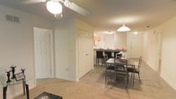 This single-floor residential unit offers a seamlessly connected open-concept layout that integrates the living, dining, and kitchen areas into one cohesive space. The living area features warm beige carpeting, providing a soft and comfortable feel, while the adjacent dining and kitchen zones transition to smooth hard flooring. A ceiling fan with integrated lighting anchors the living area, complementing the pendant light that hangs gracefully over the dining space. The dining area accommodates a dark-toned table and chair set, creating a defined gathering spot within the open floor plan. A built-in closet with classic raised-panel doors adds convenient storage near the entryway. The kitchen is positioned at the far end of the space, featuring light wood cabinetry and a raised breakfast bar with bar-height seating, making it ideal for casual dining and entertaining. Overhead lighting in the kitchen ensures the area is well-illuminated. The layout benefits from a practical flow between rooms, with doorways leading to additional areas of the unit. The overall ambiance is warm, understated, and functional, with neutral tones throughout that create a clean and inviting atmosphere.