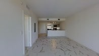 This single-floor space showcases a clean, contemporary design centered around an open-plan layout that seamlessly connects the living area to the kitchen. The flooring throughout is a striking marble-style tile in white and grey tones, lending an upscale, polished aesthetic to the entire space.

The kitchen is positioned toward the rear of the open area and features a white peninsula counter that provides both workspace and a natural divider between the living and cooking zones. Cabinetry is finished in white with a minimalist profile, and stainless steel appliances—including a refrigerator, microwave, and oven—are integrated into the layout. Recessed track lighting above the kitchen ensures the area is well illuminated.

The living area is generous and open, benefiting from a neutral white palette on the walls and ceiling that enhances the sense of brightness and space. HVAC vents are discreetly integrated into the ceiling, maintaining the clean visual lines of the interior. A hallway visible to the left leads to additional rooms, suggesting a thoughtfully organized floor plan. Overall, the space presents a bright, modern ambiance with refined finishes throughout.