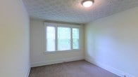 This single-floor space presents a straightforward and functional layout, highlighted by a well-lit room visible in the image. The room features a triple-panel window fitted with horizontal blinds, allowing generous natural light to filter in while framing views of lush greenery outside. The walls are finished in a soft, neutral cream tone, contributing to a calm and understated ambiance throughout.

Underfoot, wall-to-wall carpeting in a muted beige provides warmth and comfort. The ceiling draws attention with a distinctive embossed or patterned textured finish, adding a subtle decorative element to the otherwise minimalist space. A flush-mount ceiling light fixture provides overhead illumination.

Clean white baseboards line the perimeter of the room, complementing the light wall color and creating a cohesive, tidy appearance. A floor-level vent indicates a built-in climate control system. The overall feel of the space is bright, quiet, and move-in ready, suited for those seeking a simple, low-maintenance environment with natural light and modest architectural detailing.