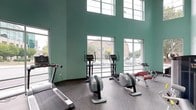 Located at 400 South Greene Street in Greenville, North Carolina, this fitness center is a well-appointed, single-floor facility designed to support a comprehensive workout experience. The space is immediately distinguished by its soaring ceilings, which lend an open, airy quality throughout the room. Signature mint-green walls add a fresh, energizing aesthetic that complements the modern design.

Abundant natural light pours in through expansive floor-to-ceiling windows and glass panels lining multiple walls, offering unobstructed views of the surrounding downtown Greenville streetscape. The generous glazing ensures the space feels bright and connected to the outdoors at all times of day.

The cardio zone is well-equipped with multiple treadmills and elliptical machines, each positioned to take full advantage of the street-level views. A stationary bike and rowing machines round out the aerobic offerings. The strength training area features a selection of weight machines, free weights, dumbbells, and flat benches, providing options for a full-body resistance workout. Decorative touches, including framed artwork and a vintage bicycle mounted high on the wall, give the space a distinctive personality. The durable speckled rubber flooring throughout is both practical and visually cohesive, completing this thoughtfully designed fitness facility.