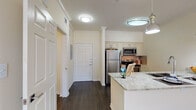 This single-floor residence is designed with a cohesive, neutral palette and functional layout that maximizes everyday comfort. The kitchen serves as a central highlight, showcasing granite countertops with a dual-basin sink, crisp white cabinetry with decorative raised-panel detailing, and a full suite of stainless steel appliances including a refrigerator, electric range, and microwave. A tile backsplash adds texture, while stylish pendant lights and a flush-mount ceiling fixture provide layered illumination over the extended kitchen island. Dark hardwood-style flooring runs throughout the kitchen and entry areas, creating a clean contrast against the warm, neutral walls accented by crown molding.

The entry hall transitions seamlessly into the open living area, where plush neutral carpeting, crown molding, and a ceiling fan with a light kit contribute to a relaxed, comfortable atmosphere. A large window allows natural light to fill the space and offers an outdoor view. The bedroom is generously proportioned with soft carpeting and its own window. The bathroom features matching granite-topped cabinetry, a tub-shower combination, and dark hardwood-style flooring, maintaining the consistent, refined aesthetic found throughout the residence.
