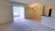 This single-floor residence offers a clean, functional layout designed for comfortable everyday living. The main living and dining area is a standout feature, showcasing plush wall-to-wall carpeting in a neutral tone that adds warmth and softness underfoot. Large sliding glass doors flood the space with abundant natural light, creating an airy, open atmosphere while providing direct access to the outdoors and views of the surrounding greenery. Vertical blinds offer flexible light control and privacy as needed. A stylish pendant light fixture is centered over the dining area, adding a touch of elegance to the open-plan layout. The walls are finished in a soft, warm beige tone that complements the overall neutral palette of the space. A floor vent ensures efficient climate control throughout the room. To one side, a partial view of a countertop hints at an adjacent kitchen area, suggesting a convenient and connected flow between living and cooking spaces. The overall ambiance is calm, bright, and welcoming, with a straightforward design that emphasizes practicality and comfort in a well-lit, open environment.