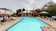 This single-story amenity facility is centered around a classic brick clubhouse building with notable curb appeal. The structure features a traditional colonial-style facade with white columns, a covered portico entrance, and a dark shingled roof accented by an arched transom window. The building is framed by mature trees and landscaping, lending a welcoming, established character to the property.

Directly in front of the clubhouse lies a generously sized outdoor swimming pool with clear turquoise water, bordered by a brick-paved deck. Lounge seating and a shaded umbrella area are positioned poolside, providing comfortable outdoor relaxation space.

Inside, the fitness center is divided into functional zones. One area houses strength-training equipment, including resistance machines and weight benches, positioned along neutral-toned walls with white wainscoting and natural light filtering through multiple windows. A separate open exercise area features rubber floor mats, a step platform, and full-length mirrored glass panels that enhance the sense of space and allow users to monitor their form. Cardio equipment, including a treadmill and an elliptical machine, is also available. A wall-mounted television adds convenience for users during workouts. The fitness center maintains a clean, practical aesthetic throughout.