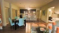 This single-floor space showcases a seamless open-concept layout that connects the kitchen, dining, and living areas into one cohesive and functional environment. Rich hardwood flooring flows throughout, lending warmth and continuity to the space.

The kitchen is a standout feature, appointed with dark espresso cabinetry, stainless steel appliances including a refrigerator and range, and a light-toned countertop island with bar seating. Pendant lighting hangs above the island, providing both task illumination and a stylish focal point. A track lighting system overhead further enhances the kitchen's functionality.

Adjacent to the kitchen, the dining area comfortably accommodates a dark wood table surrounded by distinctive teal upholstered chairs, adding a pop of color and character to the space. The living area is furnished with neutral-toned seating and accented with a patterned area rug, creating a relaxed and welcoming atmosphere.

Multiple doorways visible from the main area suggest additional rooms beyond, offering privacy and separation when needed. Decorative wall art and thoughtful furnishings contribute to the overall modern aesthetic. The open layout promotes a sense of spaciousness and allows for easy movement and interaction between the kitchen, dining, and living zones.