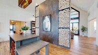 This single-floor space combines striking design elements with versatile, functional areas. The central gathering space commands attention with soaring ceilings and a dramatic floor-to-ceiling accent wall composed of stacked log cross-sections framing a textured metal panel, with curated artwork displayed at its center. Warm-toned hardwood flooring runs throughout, unifying the open layout. A comfortable lounge area features ample natural light and direct sightlines to adjacent spaces, fostering an open, connected atmosphere.

A dedicated library nook flanks the accent wall, outfitted with tall, dark-framed bookshelves, a round table, and soft seating—ideal for quiet reading or small gatherings. The kitchen is well-equipped with stainless steel appliances, a generous island, crisp white shaker cabinetry, quartz countertops, and a distinctive geometric-patterned tile backsplash that adds visual interest.

The fitness center occupies its own wing, finished in deep navy tones with large windows providing abundant natural light and outdoor views. It houses treadmills, ellipticals, stationary bikes, cable machines, free weights, and benches. A retractable glass garage door opens the weight area directly to the outdoors, enhancing ventilation and connectivity to the exterior.