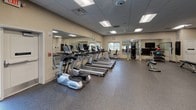This fitness center is thoughtfully designed to support a comprehensive workout experience across a single floor. The main exercise area is spacious and open, featuring durable rubber flooring and a drop ceiling with recessed panel lighting throughout. A row of True-brand treadmills lines the center of the room, complemented by elliptical trainers and a multi-station cable machine for strength training. Full-length mirrors along one wall visually expand the space and allow users to monitor their form during workouts. Natural light filters in through multiple windows, and a ceiling fan aids in ventilation and comfort.

A dedicated studio room provides additional training versatility, outfitted with a flat-screen television, a whiteboard, a storage shelving unit, and a rack stocked with medicine balls and exercise balls in various sizes. Adjustable workout benches are positioned on the rubber floor, making the room ideal for group classes or personal training sessions.

Adjacent to the main fitness area is a glass-enclosed room with frosted panel doors, visible through the corridor. The space features carpeted flooring and seating, suggesting use as a meeting or wellness consultation room. Safety features such as a wall-mounted fire extinguisher and exit signage are prominently placed. The overall environment is clean, functional, and professionally appointed.