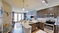 This well-appointed 1-bedroom, 1-bathroom residence is designed with a contemporary open-concept floor plan that seamlessly connects the kitchen, dining, and living areas into one cohesive living space. The kitchen is a standout feature, boasting rich dark-espresso cabinetry, granite countertops, a subway tile backsplash, and stainless steel appliances including a range and built-in microwave. A peninsula with an undermount sink and pendant lighting above provides both additional prep space and a natural gathering spot. The flooring throughout transitions between warm hardwood-style planks and complementary area rugs, adding texture and visual interest. The living area is bathed in natural light, thanks to a set of elegant French doors that open to the outside, framed by floor-length curtains. Soft blue accent walls create a calming, modern atmosphere, while recessed and track lighting provide versatile illumination throughout. The overall ambiance is clean, bright, and modern, reflecting thoughtful design choices that balance both style and functionality — making this a comfortable and visually appealing single-floor residence.