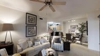Located at 101 Crossing Dr in Millbrook, Alabama, this single-floor residence offers a thoughtfully designed open-concept layout that seamlessly connects the main living areas. The living room is a welcoming focal point, featuring vaulted ceilings accented by a ceiling fan, neutral tones, and comfortable seating arranged around a round coffee table. Framed wall art adds a refined decorative touch, contributing to the space's polished ambiance. The kitchen is situated just beyond the living area in an open-plan arrangement, showcasing light wood cabinetry, stainless steel appliances including a built-in microwave, and a functional layout ideal for everyday use. A dining area flows naturally from the kitchen, featuring a dark wicker dining set positioned beneath track lighting mounted on a tiled accent wall — a distinctive architectural detail that adds character and texture to the space. The continuous flooring and open sightlines throughout create a cohesive, airy feel. The overall aesthetic balances casual comfort with tasteful design, making the space both functional and inviting. The single-story layout ensures convenient accessibility, while the well-considered interior design elements give the home a warm, livable character.