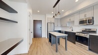 This well-appointed single-floor residence showcases a contemporary design with clean lines, light hardwood flooring, and a bright, neutral color palette throughout. The kitchen serves as the centerpiece of the home, featuring a striking two-tone cabinetry scheme with light gray upper cabinets and dark lower cabinets, a herringbone-patterned tile backsplash, white quartz countertops, and a full suite of stainless steel appliances including a gas range, microwave, dishwasher, and refrigerator. A central island with open shelving and pendant lighting suspended from an exposed wood beam adds both function and character to the space. The bedrooms are generously sized, with large floor-to-ceiling windows that invite abundant natural light and offer expansive urban views of the surrounding cityscape. One bedroom features soft carpeting, while the other benefits from the same hardwood flooring found throughout the main living areas. The bathroom is finished in a clean, modern style with white shaker cabinetry, a framed mirror, matte black fixtures, and a tiled tub-shower combination. A dedicated utility area includes a stacked washer and dryer alongside built-in shelving and a walk-in closet, providing excellent storage and convenience. Recessed lighting throughout ensures a consistently well-lit environment across all areas of the home.
