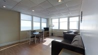 This single-floor space offers a welcoming open-concept layout that seamlessly combines a living area and dining area into one cohesive, light-filled environment. Large corner windows wrap around the space, flooding the interior with natural light and framing sweeping panoramic water views that serve as a stunning backdrop throughout the day. The drop-panel ceiling with recessed lighting complements the neutral, warm-toned walls to create a clean and understated aesthetic. Hardwood-style flooring runs throughout, adding warmth and continuity to the space. The dining area features a compact round table with mixed seating, making it an intimate spot for meals while enjoying the surrounding views. A media console with a flat-screen television anchors one wall, providing a functional entertainment setup within the living zone. The living area is furnished with a comfortable dark upholstered sofa set arranged to maximize both comfort and the striking waterfront sightlines. A floor lamp adds a soft ambient touch to the seating area. Baseboard heating units are visible along the windowed walls, ensuring year-round comfort. The overall ambiance is relaxed and airy, with the expansive windows and elevated vantage point offering an immersive connection to the outdoors.