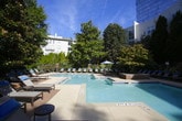 This outdoor amenity space centers around a generously sized swimming pool with clear, turquoise water, clearly marked with depth indicators and a designated no-diving zone for safety. The pool is complemented by a separate wading or splash area, making it suitable for a range of users. Surrounding the pool, a wide concrete and brick-paved sundeck provides ample room for relaxation, furnished with rows of cushioned lounge chairs arranged to take full advantage of the open sky and natural light.

Mature trees and well-maintained landscaping line the perimeter of the pool area, offering natural shade and a sense of privacy. The greenery creates a pleasant, park-like atmosphere that softens the urban setting. Additional seating areas with tables and chairs are thoughtfully positioned around the deck, ideal for socializing or casual outdoor dining.

Access to the pool area is controlled through a secure wrought-iron gate framed by classic brick pillars, ensuring a private and safe environment. The surrounding multi-story building features balconies that overlook the courtyard, integrating the outdoor space seamlessly with the residential structure. Overall, this outdoor space is designed to provide a comfortable, resort-like retreat with practical amenities and inviting landscaping.