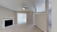 This single-floor residence offers a thoughtfully designed layout with a neutral color palette and comfortable finishes throughout. The spacious living room is a standout, featuring plush carpeting, a classic white-mantled fireplace, a ceiling fan, and a large sliding glass door dressed with vertical blinds that invites natural light into the space. The vaulted ceiling adds an airy, open feel to the living and adjacent dining area, which connects seamlessly to the kitchen via a breakfast bar with light stone-look countertops. The galley-style kitchen is well-equipped with dark espresso cabinetry, light granite-style countertops, wood-look plank flooring, and a full suite of black appliances including a range, microwave, refrigerator, and dishwasher. A skylight fixture keeps the kitchen bright and functional. The bathroom features a dark wood vanity with a stone-look countertop, a wall-mounted medicine cabinet, and direct access to a generous walk-in closet complete with built-in shelving and carpeted flooring. Throughout the home, consistent warm-gray tones, textured ceilings, and clean white trim create a cohesive and inviting ambiance.