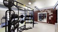 This fitness center offers a thoughtfully designed workout space that combines functionality with a polished aesthetic. The single-level layout maximizes usable floor space, accommodating a wide variety of training styles. A central multi-functional rig serves as the focal point, outfitted with TRX suspension trainers and battle ropes for functional and high-intensity training. Surrounding the rig, neatly organized racks hold an extensive selection of dumbbells, kettlebells, medicine balls, and weight plates, ensuring accessibility for all fitness levels. A dedicated squat and power rack station is visible toward the rear, providing space for barbell-based strength training. Cardio equipment, including treadmills, can be seen toward the back of the space near large windows that invite natural light. The interior features warm wood-paneled accent walls that contrast with the light-toned flooring and white ceilings, creating a balanced and motivating environment. Pendant and linear LED lighting illuminate the space evenly. Full-length mirrors along the walls enhance the sense of openness while allowing users to monitor their form throughout their workouts.