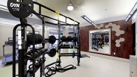 This fitness center offers a thoughtfully designed workout space that combines functionality with a polished aesthetic. The single-level layout maximizes usable floor space, accommodating a wide variety of training styles. A central multi-functional rig serves as the focal point, outfitted with TRX suspension trainers and battle ropes for functional and high-intensity training. Surrounding the rig, neatly organized racks hold an extensive selection of dumbbells, kettlebells, medicine balls, and weight plates, ensuring accessibility for all fitness levels. A dedicated squat and power rack station is visible toward the rear, providing space for barbell-based strength training. Cardio equipment, including treadmills, can be seen toward the back of the space near large windows that invite natural light. The interior features warm wood-paneled accent walls that contrast with the light-toned flooring and white ceilings, creating a balanced and motivating environment. Pendant and linear LED lighting illuminate the space evenly. Full-length mirrors along the walls enhance the sense of openness while allowing users to monitor their form throughout their workouts.