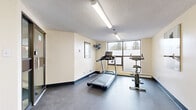 Located at 2710 Saratoga Pl in Ottawa, Ontario, this fitness centre offers a dedicated space for residents to maintain an active lifestyle. The room features a practical, no-frills layout with durable dark rubber flooring designed to withstand heavy equipment use and high-traffic activity.

The cardio area is equipped with a treadmill and a stationary exercise bike, both positioned near large windows that allow natural light to fill the space and offer views of the surrounding trees — creating an pleasant environment for workouts. A wall-mounted fan provides additional air circulation in this area.

On the opposite side of the room, a multi-station cable weight machine provides options for strength training exercises. Motivational fitness photography is displayed on the walls, adding an energizing aesthetic to the space. A posted Fitness Centre Rules sign ensures the facility is used responsibly by all.

The room is illuminated by fluorescent strip lighting mounted to a textured ceiling, providing bright and even coverage throughout. Entry is through a set of glass-paneled doors, offering visibility into the space from the adjoining corridor. The walls are finished in a neutral cream tone, keeping the environment clean and functional. This fitness centre serves as a convenient, well-equipped amenity for building residents.