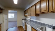 This single-floor, one-bedroom, one-bathroom residence offers a thoughtfully designed layout that balances functionality and comfort. The kitchen is a standout feature, equipped with warm medium-toned wooden cabinetry that extends to the upper walls, providing ample storage. Granite-style countertops in a speckled grey-blue tone add a durable and attractive work surface, complemented by white appliances including a gas range and dishwasher. A skylight overhead floods the kitchen with natural light, creating a bright and welcoming cooking environment. The kitchen opens directly into the living area, which features rich hardwood-style flooring that extends throughout the main living spaces, adding warmth and visual continuity. A window in the living area allows additional natural light to filter in. Neutral wall tones throughout the residence create a versatile backdrop suitable for a variety of décor styles. The overall layout is efficient and practical, making excellent use of its generous square footage to deliver a comfortable single-floor living experience.