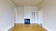 This single-floor space combines classic architectural details with a clean, neutral palette to create a warm and inviting environment. Hardwood floors run throughout, lending warmth and continuity from room to room. The main living area is a standout, anchored by a decorative brick fireplace framed by a white mantel — a charming focal point that adds character and a sense of history to the space. Adjacent to the fireplace, a built-in shelving nook provides practical storage while enhancing the room's traditional aesthetic. Crown molding traces the perimeter of the ceiling, adding a refined architectural touch that elevates the overall feel of the space. The walls are finished in a soft, neutral gray tone that complements the natural wood floors and white trim throughout. Doorways with classic white framing connect the rooms in a straightforward, functional layout, allowing for easy flow between spaces. The corridor view reveals the generous proportions of the layout, with multiple rooms extending beyond. Overall, the space carries a timeless residential character, ideal for those who appreciate classic craftsmanship and understated elegance.