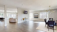 Located in Marietta, Georgia, this thoughtfully designed single-floor community clubhouse offers a generous, open-plan layout that seamlessly connects multiple functional zones. Rich hardwood-style flooring runs throughout, tying together the space's contemporary aesthetic defined by neutral gray tones, white trim, and abundant natural light streaming through oversized windows.

The main lounge area provides a relaxed gathering space, complemented by recessed lighting, a wall-mounted television, and curated abstract artwork. Large picture windows frame views of the surrounding mature trees, creating a bright and inviting ambiance. An adjoining dining zone features an elegant gold-toned chandelier and ample seating for group gatherings.

The kitchen is a standout feature, showcasing crisp white shaker cabinetry with brass hardware, a striking herringbone tile backsplash, stainless steel appliances, and a boldly accented blue island with a quartz countertop. A secondary countertop wall with additional cabinetry and storage provides further functionality.

A distinctive bar and lounge area includes high-top seating and floor-to-ceiling glass garage doors that open directly onto an outdoor pool deck, blurring the line between interior and exterior. This indoor-outdoor connection enhances the versatility of the space, making it well-suited for both casual everyday use and larger social events.