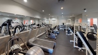 This single-floor fitness facility is thoughtfully designed to support a full range of workout routines. The main exercise area features durable dark rubber flooring throughout, providing both safety and practicality underfoot. Recessed lighting illuminates the space evenly, complemented by natural light filtering in through multiple windows positioned along the walls.

The cardio zone is well-stocked with treadmills, upright and recumbent stationary bikes, and elliptical machines, all arranged along a wall-length mirror that visually expands the space and allows users to monitor their form. The strength-training area features an array of white cable and weight machines, a bench press station, and additional resistance equipment, offering versatile options for full-body workouts. Wall-mounted televisions are positioned throughout for convenience.

Multiple exit doors enhance accessibility and safety throughout the facility. Adjacent to the main fitness floor is a dedicated children's play room, featuring vibrant orange and black painted walls accented by a decorative border, carpeted flooring, and an assortment of toys and child-sized furnishings. Two windows provide natural light, creating a cheerful and engaging environment for younger visitors while caregivers exercise nearby.