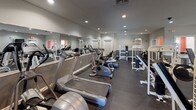 This single-floor fitness facility is thoughtfully designed to support a full range of workout routines. The main exercise area features durable dark rubber flooring throughout, providing both safety and practicality underfoot. Recessed lighting illuminates the space evenly, complemented by natural light filtering in through multiple windows positioned along the walls.

The cardio zone is well-stocked with treadmills, upright and recumbent stationary bikes, and elliptical machines, all arranged along a wall-length mirror that visually expands the space and allows users to monitor their form. The strength-training area features an array of white cable and weight machines, a bench press station, and additional resistance equipment, offering versatile options for full-body workouts. Wall-mounted televisions are positioned throughout for convenience.

Multiple exit doors enhance accessibility and safety throughout the facility. Adjacent to the main fitness floor is a dedicated children's play room, featuring vibrant orange and black painted walls accented by a decorative border, carpeted flooring, and an assortment of toys and child-sized furnishings. Two windows provide natural light, creating a cheerful and engaging environment for younger visitors while caregivers exercise nearby.