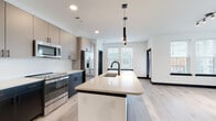 This contemporary single-floor residence showcases a thoughtfully designed open-concept layout with a clean, modern aesthetic throughout. The kitchen is a standout centerpiece, featuring a generous island with a light quartz countertop, an undermount sink with a matte black faucet, and sleek pendant lighting overhead. Cabinetry is finished in a two-tone palette of warm taupe uppers and dark lower cabinets, complemented by a classic white subway tile backsplash. Stainless steel appliances, including a French door refrigerator, range, and built-in microwave, complete the well-appointed culinary space.

Light-toned hardwood flooring flows seamlessly throughout the residence, accented by bold dark baseboards and dark-framed interior doors that lend a sophisticated contrast. The bedrooms are bright and airy, benefiting from multiple windows that invite abundant natural light and views of the surrounding neighborhood. One bedroom includes a walk-in closet for added convenience.

The bathrooms reflect the same modern sensibility, with one featuring a soaking tub surrounded by floor-to-ceiling subway tile and matte black fixtures, and another offering a walk-in glass shower alongside a wood-toned vanity with quartz countertops and a large framed mirror. Recessed lighting throughout ensures a bright, welcoming ambiance in every room.