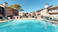 The second pool area is a well-appointed outdoor amenity space designed for relaxation and recreation. At its center is a generously sized swimming pool with clear, blue-tinted water, framed by a textured, aggregate concrete deck that provides comfortable, slip-resistant footing. A shallow wading area with brick tile edging adjoins the main pool, offering a gentler entry point for swimmers of all levels, with depth markers clearly posted for safety.

Adjacent to the pool, a dedicated spa area is enclosed by a low brick wall and governed by posted spa policies, ensuring a regulated and enjoyable experience for residents and their guests. The deck is furnished with teal lounge chairs and multiple high-top dining tables paired with matching teal chairs, each shaded by vibrant green market umbrellas — ideal for outdoor dining or socializing in the sun.

The area is fully enclosed by an iron security fence with gated access, providing both safety and privacy. Mature trees displaying rich autumn foliage surround the perimeter, offering natural shade and a scenic backdrop. Multi-story brick residential buildings frame the space, lending it a cohesive, community-oriented character. Abundant natural light fills the area throughout the day, creating a bright and inviting outdoor retreat.