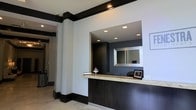 This single-floor space presents a contemporary reception area designed with a sharp contrast of dark and light elements. The front desk serves as the focal point, featuring a light granite or stone countertop paired with a dark wood base, offering both functionality and visual appeal. Behind the desk, a large rectangular mirror is mounted against a dark accent wall, enhancing the sense of depth within the space. Recessed ceiling lights are strategically positioned to illuminate the desk area and surrounding walls, providing a clean, well-lit environment. Bold dark trim lines the base of the walls and the ceiling perimeter, creating a defined architectural framework against the white walls. The flooring appears to be light-toned tile, complementing the overall color palette. A framed photo and decorative glass dish accessorize the desk, adding subtle warmth to the otherwise minimalist design. A corridor visible in the background suggests additional rooms or offices beyond the reception zone. The overall ambiance is professional, modern, and welcoming, well-suited for a business, medical, or hospitality environment.