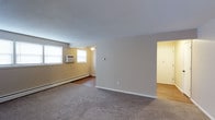 This single-floor space features an open-concept living area that combines functionality with a straightforward layout. The main living area is finished with wall-to-wall carpeting, while an adjacent section transitions to hardwood-style flooring, providing a clear visual distinction between zones. Two windows along the exterior wall allow natural light to brighten the space, complementing the warm, neutral-toned walls throughout.

Climate control is handled by a wall-mounted air conditioning unit positioned beneath the windows, alongside baseboard heating that runs along the perimeter of the room — offering year-round comfort. The ceiling is smooth and white, featuring a centrally placed ceiling light fixture that provides general illumination.

A doorway visible on the right side of the space leads to an adjoining room or hallway, suggesting additional areas beyond the main living space. The white baseboards and clean wall lines contribute to a tidy, minimalist aesthetic. Overall, the space presents a practical and functional layout suited for everyday living, with essential utilities already in place.