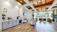 Located at 400 Hidden Palm Cir in Kissimmee, Florida, this single-floor community space is a beautifully appointed clubhouse and leasing center that blends coastal elegance with modern functionality. The interior is unified by rich, wide-plank hardwood flooring that runs throughout, complemented by a crisp white palette and warm wood-paneled coffered ceilings that lend both height and architectural character to the space.

The welcoming reception area features a natural wood communal table, woven accent chairs, and a prominent reception desk, all bathed in natural light streaming through large glass entry doors. A dedicated service counter with white shaker cabinetry, quartz countertops, and white shiplap tile backsplash provides a refreshment station for guests and residents alike.

The expansive living area is furnished with neutral linen seating, woven rattan accent chairs, and layered rugs that create a relaxed yet refined atmosphere. A textured wall panel anchors a wall-mounted flat-screen television, while lush indoor plants add a vibrant natural touch. Dramatic floor-to-ceiling windows along one full wall frame sweeping outdoor views of a resort-style pool and swaying palm trees, flooding the space with sunlight. Statement drum chandeliers with warm metallic finishes complete the sophisticated, resort-inspired ambiance throughout.