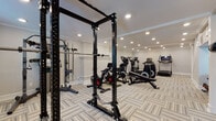 The fitness center at Montrose Brookhaven in Brookhaven, Georgia is a thoughtfully designed, single-floor facility that provides residents with a comprehensive workout environment. The space is finished with neutral gray walls, elegant crown molding, and a herringbone-patterned carpet tile flooring throughout, creating a clean and polished aesthetic.

The facility is organized across interconnected zones to accommodate a variety of training styles. The cardio area is well-stocked with multiple stationary bikes, elliptical trainers, and treadmills, each equipped with individual screens. Adjacent to this, a dedicated strength training zone features a full power rack with barbell and weight plates, along with a weight bench and cable attachments for versatile resistance training.

A separate functional training room provides open floor space ideal for dynamic workouts. This area includes resistance bands, a suspension trainer, a plyo box, medicine balls, and a full rack of dumbbells in varying weights, catering to both beginner and advanced fitness routines. Wall-mounted mirrors enhance the sense of space and allow for form checking during exercises.

Recessed ceiling lighting illuminates the entire facility brightly and evenly, while windows allow natural light to supplement the interior lighting. Posted gym rules and informational signage contribute to a well-organized, community-focused atmosphere throughout the space.