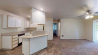 This single-floor unit is organized around a practical, open layout that connects the kitchen and living areas seamlessly. The kitchen is well-equipped with white cabinetry featuring light wood trim, a built-up overhead cabinet structure housing the range hood, a white refrigerator, a dishwasher, and a gas cooktop. A peninsula with a light-colored laminate countertop extends into the space, providing additional prep area and serving as a natural divider between the kitchen and the rest of the unit. Tile flooring with a warm stone-like pattern runs throughout the kitchen and entry areas.

The living area is generously sized and carpeted in a neutral taupe tone, offering a comfortable transition from the hard flooring of the kitchen. A ceiling fan with an integrated light fixture is centrally positioned, and vertical blinds on one wall suggest access to natural light. The room provides ample wall space and multiple electrical outlets.

The bedroom is a well-proportioned, carpeted room with neutral walls and multiple outlets, providing a quiet and versatile space. The bathroom features the same warm-toned tile flooring found in the kitchen, a single-sink vanity with light wood cabinetry, a toilet, and a full tub-shower combination with chrome fixtures and a curtain rod.