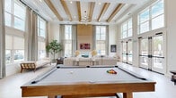 Located at 8060 Purchase Pl NE in Leland, NC, this impressive single-floor community clubhouse spans a generously sized footprint designed to serve as a premier social and recreational hub. The centerpiece is a grand living room distinguished by soaring ceilings with exposed wood beam detailing, a classic fireplace, a wall-mounted TV, and abundant natural light streaming through oversized double-height windows and French doors. A full-size billiards table anchors the space, complementing the plush sectional seating arranged around a coastal-inspired area rug. The welcoming entryway features warm vertical shiplap wood paneling, light hardwood flooring, a statement round pedestal table, and a steel-framed sliding barn door. A combined dining and lounge area offers herringbone-tiled flooring, cozy mixed seating, and views of the outdoor pool area. The kitchen is finished with crisp white tile backsplash, light stone countertops, a stainless steel sink, and open wood shelving with brass hardware accents. The dedicated exercise room is equipped with multiple treadmills and cardio machines, light wood flooring, mirrored walls, and a ceiling fan. Outdoors, a large resort-style swimming pool is surrounded by a spacious sun deck with lounge chairs, set against a backdrop of lush Carolina pines.
