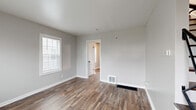 Located at 4470 E Broad St in Columbus, Ohio, this multi-floor home offers two bedrooms, one bathroom, and a full basement spread across three levels. The main floor welcomes you with a bright living room featuring rich, wood-grain luxury vinyl flooring, neutral gray walls, and a front entry door flanked by windows that fill the space with natural light. A staircase with a sleek metal railing leads to the upper levels. The kitchen is a standout, outfitted with dark shaker-style cabinetry, white countertops, a stainless-style sink, and a full suite of black appliances including a gas range, dishwasher, and refrigerator. An adjacent dining area includes a ceiling fan and additional windows. The bathroom features a full tub-shower combo with white tile surround, a natural light window, and wood-grain flooring. Both bedrooms are generously sized with neutral tones, hardwood or wood-style flooring, and ample natural light from multiple windows. The primary bedroom is a comfortable size, while the second bedroom features a distinctive textured ceiling detail. The unfinished basement provides additional storage or utility space with painted concrete block walls and visible ductwork overhead.