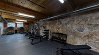 This fitness space offers a distinctive workout environment that blends industrial character with practical functionality. The space is defined by its striking exposed stone walls, which carry a weathered, textured appearance that speaks to the building's historic origins. Overhead, warm wood-plank ceiling panels run the length of the room, complemented by exposed ductwork, piping, and fluorescent strip lighting that reinforces the raw, utilitarian aesthetic.

The flooring throughout is covered in durable rubber gym matting, providing cushioning and traction across the entire workout area. The space is well-equipped for a full-body fitness regimen. Cardio equipment includes multiple elliptical trainers, stationary bikes, and a NordicTrack commercial treadmill. For strength training, the facility features multiple dumbbell racks with a complete range of weights, kettlebell sets, adjustable workout benches, and a barbell rack with a squat/press station. Additional accessories such as resistance equipment and storage shelving are also present.

Natural light filters in through windows at one end of the space, offering a subtle connection to the outdoors. A secured entry door with keypad access provides controlled entry. The combination of historic architectural elements and modern fitness equipment creates a focused, motivating atmosphere that is both functional and visually engaging.