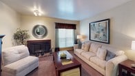 This single-story residence offers a well-organized layout encompassing a living room, dining area, kitchen, multiple bedrooms, and two full bathrooms. The living room serves as a central gathering space, featuring a wood-burning fireplace with brick surround, carpet flooring, a textured ceiling, and a window with decorative drapery that allows natural light to filter in. Adjacent to the living room, the dining area accommodates a glass-top table with seating for four, pendant lighting overhead, and tile flooring that transitions from the nearby kitchen. The kitchen is equipped with warm wood cabinetry, tile flooring, a full-size refrigerator, and a range, providing functional storage and workspace. The bedrooms feature carpet flooring, ceiling fan light fixtures, and neutral wall tones. One bedroom includes a walk-in closet, while another benefits from a window with blinds. Both bathrooms share a consistent design, each offering a single-sink vanity with wood cabinetry, a large mirror with overhead lighting, and a tub-shower combination with a decorative curtain. Tile flooring completes each bathroom. Throughout, the residence maintains a cohesive warm and neutral palette.