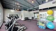 This fitness center offers a comprehensive workout environment designed to accommodate a wide range of exercise routines. The space features an open, industrial-style ceiling with exposed ductwork and track lighting, lending a modern, energetic atmosphere throughout. The durable purple rubber flooring provides both comfort and functionality underfoot, ideal for high-intensity training sessions.

The cardio zone is lined with elliptical machines and other aerobic equipment positioned along one side of the room, allowing ample space to move freely. A dedicated free weights area includes a multi-tiered dumbbell rack alongside an adjustable workout bench, catering to strength training needs at various levels. Full-length mirrors along the walls enhance the sense of space while allowing users to monitor their form during exercise.

A well-organized functional training rack along one wall stores a variety of medicine balls, resistance bands, and exercise mats, offering versatile options for dynamic and bodyweight workouts. Natural light filters in through windows visible at the far end of the space, contributing to a bright and motivating environment. The overall layout is thoughtfully arranged to maximize usability while maintaining an open, uncluttered feel throughout the facility.