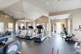 Located at 10209 Baltimore Ave in College Park, Maryland, this well-appointed fitness center offers a generous, open floor plan designed to accommodate a comprehensive workout experience. The space is defined by its striking vaulted ceilings, which rise dramatically overhead and are complemented by multiple ceiling fans that ensure comfortable air circulation throughout. Elegant white decorative columns are positioned throughout the room, adding a refined architectural character to the functional space.

The fitness center is outfitted with a wide array of equipment across dedicated zones. The cardio area features multiple treadmills, elliptical trainers, and stationary bikes, including recumbent models, providing options for varied aerobic workouts. A strength and functional training section includes a multi-station cable machine, a weight bench with rack, stability balls, and resistance accessories, catering to a full range of strength-training needs.

Large, white-trimmed windows line the walls, flooding the interior with natural light and offering views of the surrounding outdoor environment. The durable rubberized flooring throughout provides both safety and comfort underfoot. Full-length mirrors on one wall enhance the sense of space while serving a practical purpose for form monitoring. A set of glass-paned French doors provides an inviting entryway, reinforcing the bright and welcoming ambiance of this well-equipped facility.