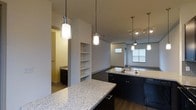 Located in Richardson, this contemporary multi-floor space spans three levels and showcases a thoughtfully designed interior with modern finishes throughout. The kitchen serves as a central highlight, featuring expansive granite countertops with a speckled finish, dark cabinetry, and a functional island with a built-in sink. Elegant pendant lights hang above the counters, providing focused task lighting while adding a refined aesthetic touch. Track lighting in the adjoining area further enhances the ambiance. The open-concept layout connects the kitchen seamlessly to the living area, which benefits from natural light streaming through large windows and is outfitted with a ceiling fan for added comfort. Built-in shelving integrated into the wall provides practical storage while contributing to the architectural character of the space. Rich hardwood-style flooring runs throughout, offering a warm contrast to the neutral wall tones. A glimpse into an adjacent room reveals dark cabinetry and tile flooring, suggesting a well-appointed bathroom or utility space nearby. The overall design balances functionality with contemporary style, making it a well-structured and visually cohesive environment across all three floors.