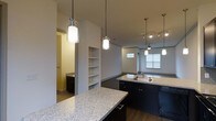 Located in Richardson, this contemporary multi-floor space spans three levels and showcases a thoughtfully designed interior with modern finishes throughout. The kitchen serves as a central highlight, featuring expansive granite countertops with a speckled finish, dark cabinetry, and a functional island with a built-in sink. Elegant pendant lights hang above the counters, providing focused task lighting while adding a refined aesthetic touch. Track lighting in the adjoining area further enhances the ambiance. The open-concept layout connects the kitchen seamlessly to the living area, which benefits from natural light streaming through large windows and is outfitted with a ceiling fan for added comfort. Built-in shelving integrated into the wall provides practical storage while contributing to the architectural character of the space. Rich hardwood-style flooring runs throughout, offering a warm contrast to the neutral wall tones. A glimpse into an adjacent room reveals dark cabinetry and tile flooring, suggesting a well-appointed bathroom or utility space nearby. The overall design balances functionality with contemporary style, making it a well-structured and visually cohesive environment across all three floors.