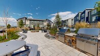 Located at 1890 S Quebec Way in Denver, Colorado, this outdoor communal space serves as a central gathering hub within a contemporary multi-unit residential community. The area is thoughtfully designed with a spacious stone-paver courtyard that provides a clean, durable foundation throughout the space.

A standout feature is the built-in outdoor kitchen, constructed with a stacked stone base and granite countertop, equipped with stainless steel grills — ideal for community gatherings and al fresco dining. Adjacent to the kitchen, Adirondack-style chairs and round picnic tables are positioned throughout the patio, offering multiple casual seating options.

A circular fire pit area anchors the center of the courtyard, surrounded by additional seating and bordered by natural landscaping elements including ornamental grasses, young deciduous trees, and decorative rock arrangements. These plantings add texture and seasonal color while creating a sense of separation between activity zones.

The surrounding multi-story residential buildings feature modern architectural styling with bold blue and gray siding, stone accents, and private balconies. The open layout allows for abundant natural light, and the Colorado sky provides a bright, expansive backdrop. The overall design balances functional amenity programming with inviting, community-centered outdoor living.