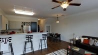 This two-story residence showcases an open-concept main living area where the kitchen, dining, and living spaces flow seamlessly into one another. Dark hardwood flooring runs throughout, providing a rich contrast against the light-colored walls and white trim. The kitchen features a breakfast bar with seating for multiple guests, granite-style countertops, and stainless steel appliances, including a full-size refrigerator. A wine rack adds a functional decorative touch above the kitchen cabinetry. The dining area accommodates a full table and chair set, positioned conveniently between the kitchen and living space. Overhead, a ceiling fan with an integrated light fixture anchors the main living area, while pendant lighting and track lighting provide additional illumination throughout the open space. Crown molding details the upper walls, adding a refined architectural touch. A hallway visible in the background suggests additional rooms and access to the second floor, extending the overall functionality of this well-appointed, multi-level residence.