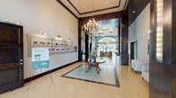 This upscale lobby and leasing center is defined by its grand, open layout and refined architectural details. The main entrance foyer makes an immediate impression with soaring ceilings adorned with crown molding, a striking multi-arm chandelier, and polished travertine tile flooring inlaid with a decorative mosaic border in rich dark marble. A central round table with artful ceramic accents serves as the focal point beneath the chandelier, with large glass entry doors allowing natural light and outdoor views to pour in from the rear. Dark espresso-toned wood columns and pilasters frame the space, creating a sophisticated contrast against the light walls and floors. Backlit accent panels between the columns add a warm, ambient glow throughout. The corridor leading deeper into the building is flanked by these illuminated columns and lush potted topiaries, guiding visitors seamlessly through the space. An adjacent lounge area features floor-to-ceiling drapery, oversized windows overlooking an outdoor courtyard, plush seating, and a built-in dark wood credenza with curated artwork. A separate business or leasing office offers a comfortable and functional setting with carpeted flooring, built-in dark wood shelving, task seating, and ample natural light. Together, these areas create a welcoming, hotel-inspired atmosphere that is both polished and approachable.