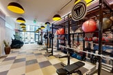 This expansive fitness center offers a thoughtfully designed, multi-zone workout facility spread across a single floor. The space is defined by its striking checkerboard-patterned flooring in neutral tones of tan, beige, and brown, which runs throughout and lends a bold, cohesive aesthetic. Overhead, dramatic black pendant lights with vibrant yellow interiors provide warm, focused illumination, complemented by recessed cove lighting with a pink-purple hue that traces the ceiling perimeter in the main workout hall.

The primary gym floor is generously proportioned, housing a robust strength training area with power racks, barbells, weight plates, and organized storage for medicine balls and resistance equipment — all set against a large-scale, expressionist-style mural that serves as a dramatic focal point. A row of premium cardio machines, including ellipticals, stationary bikes, and treadmills, lines the perimeter near a floor-to-ceiling glass wall that invites natural light and exterior views.

A secondary room, finished in warm earthy tones, functions as a dedicated studio space equipped with an interactive display screen, full-wall mirrors with ornate gold frames, stability balls, and step platforms — ideal for classes or personal training. Exposed brick columns and warm wood-framed doorways add architectural character throughout, creating an upscale, energetic fitness environment.