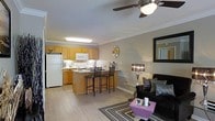 This single-floor residence offers an open-concept layout that seamlessly connects the kitchen and living area, creating a bright and functional living space. The kitchen is outfitted with warm honey-toned wood cabinetry, a decorative tile backsplash, white appliances including a full-size refrigerator and microwave, and ample counter space. A peninsula-style breakfast bar with seating for two serves as the natural dividing point between the kitchen and the main living area, making it ideal for casual dining and entertaining. The living area features light wood-style flooring that flows throughout the space, a ceiling fan with integrated lighting, and tasteful wall decor that adds character to the neutral-toned walls. Crown molding adds an elegant finishing touch along the ceiling line. The living room accommodates a comfortable seating arrangement centered around a striped area rug, with accent lighting and a decorative mirror enhancing the sense of depth and warmth. The overall ambiance is cozy yet modern, with a cohesive design aesthetic that balances practicality and style throughout the open floor plan.