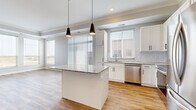 This single-floor residence offers a well-designed layout across two bedrooms, two bathrooms, a closet, and a combined kitchen and living area. The open-concept kitchen is a standout feature, boasting white shaker cabinetry with black hardware, a subway tile backsplash, granite countertops, and a central island with pendant lighting overhead. Stainless steel appliances — including a refrigerator, range, microwave, and dishwasher — complete the kitchen, which flows seamlessly into the living area via warm-toned hardwood-style flooring. Natural light filters in through multiple windows fitted with blinds throughout the common areas. The primary bedroom features hardwood-style flooring, crown molding, and a large window with outdoor views, while the second bedroom is finished with plush carpeting and a closet with panel doors. The en-suite primary bathroom is elegantly appointed with a granite-top double vanity, black-framed mirrors, wood-look flooring, and a glass-enclosed walk-in shower. The second bathroom mirrors the same refined aesthetic with a granite vanity, black-framed mirror, and a full tub-shower combination. A built-in shelving nook near the entryway adds a practical and decorative touch to this cohesive, modern residence.