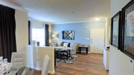 This single-floor residence in Irving, Texas, offers a well-designed, open-concept layout that promotes a sense of spaciousness and easy flow between living areas. The main living space is highlighted by warm wood-look flooring that extends throughout, providing a cohesive and inviting foundation for the entire floor plan.

The living room features soft blue accent walls that lend a calm, welcoming ambiance to the space. A comfortable seating arrangement is anchored by a patterned area rug, complemented by a sleek coffee table and a coordinating side table. A decorative mirror serves as a focal wall accent, adding depth and visual interest. Crown molding along the ceiling adds a refined architectural touch.

Natural light filters in through windows dressed with dark curtain panels, creating a pleasant contrast against the lighter wall tones. The front entry door is clearly defined, offering a clean and functional transition point into the home.

Adjacent to the living area, a dining space with bar-style seating connects seamlessly, making the layout ideal for everyday living and entertaining. The open sightlines between the kitchen, dining, and living zones reinforce the home's functional, interconnected design. Overall, this Irving residence combines practical layout with tasteful finishes for a comfortable living environment.
